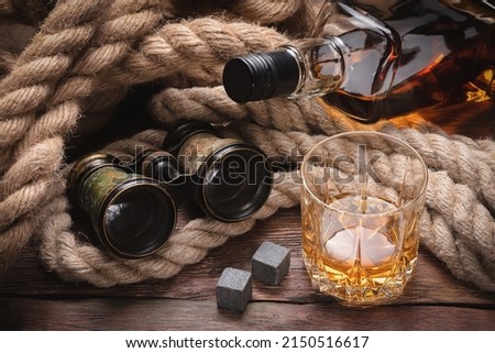 Whiskey in the drinking glass, bottle, binoculars and mooring rope on the brown wooden table background close up.