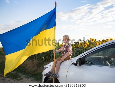 boy sits on white car with large Ukrainian flag against backdrop of blooming field of sunflowers. Independence Day Pride, freedom. patriotic education. Children against war. Ukrainians ask for peace