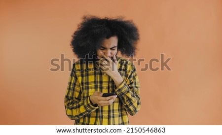 A young man with an African hairstyle on an orange background looks at the phone and is happily surprised. Emotions on a colored background
