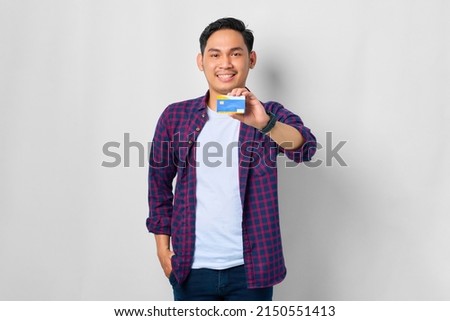 Smiling young Asian man in plaid shirt showing credit card isolated on white background