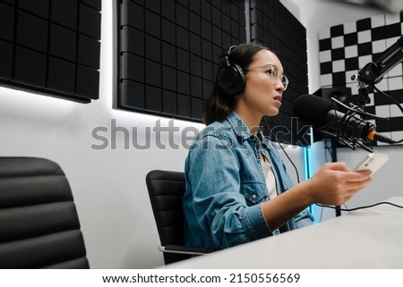 Beautiful happy young female radio host using microphone and headphones while broadcasting in studio