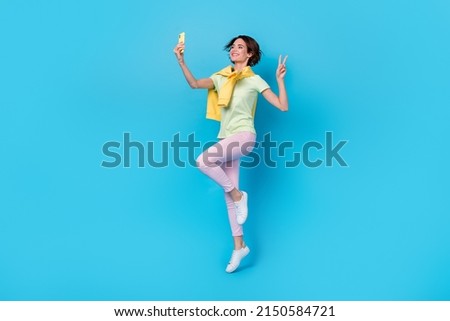 Full length photo of cute millennial brunette lady jump do selfie wear t-shirt pants shoes isolated on blue background