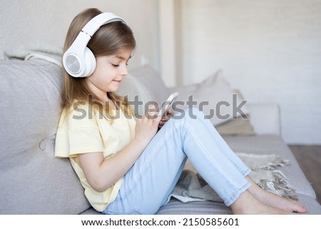 Little girl sitting on sofa with earphones and watching cartoons on mobile phone. 
