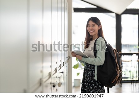 Portrait young asian woman student holding digital tablet at college standing in front of locker with backpack.