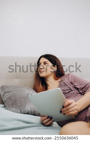 Smiling pregnant young woman lying on bed and using tablet at home. Pregnancy, technology, people and expectation concept - close up of pregnant woman with tablet pc computer in bed at home