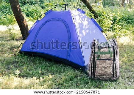 Defocus blue open tourist tent standing on green nature background. Army backpack. Tourism concept. Summer vacation in forest, camping. Blurred. Out of focus.