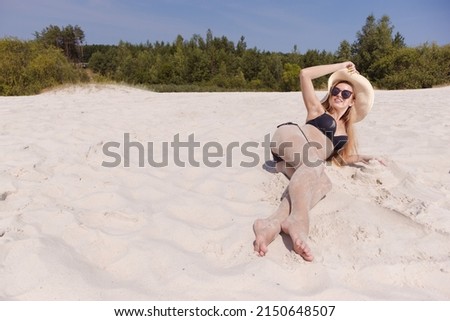 A blonde girl with long hair in a white hat is resting on the light sand near the sea on a sunny day