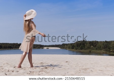 A blonde girl with long hair in a white hat is resting on the light sand near the sea on a sunny day