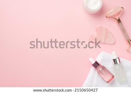 Skincare concept. Top view photo of rose quartz roller gua sha massager cream jar two glass transparent bottles and white towel on pastel pink background with blank space