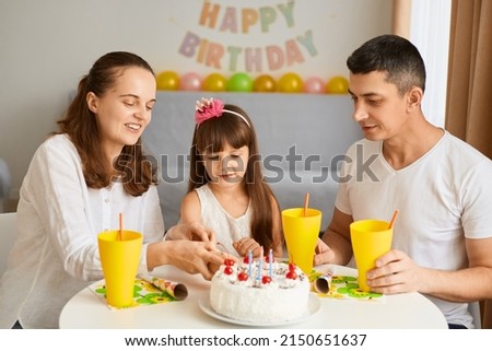 Image of happy positive family celebrating good event, sitting at table with birthday cake and drink, charming kid cutting holiday dessert together with mother, expressing good emotions.