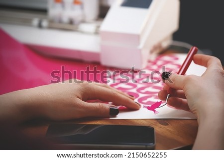 woman with painted nails weeds freshly cut peace dove stickers in pink color from carrier film. wooden worktop with plotting machine in background and mobile phone in forground. selective focus
