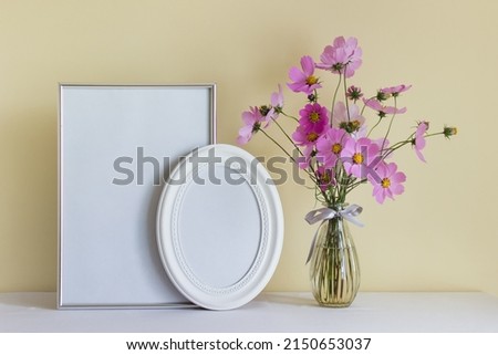 Vertical A4 frame and oval white frame mockup blank frame with pink summer flowers in glass vase on yellow wall background.