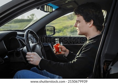 The driver, a young man driving a car, drinks an alcoholic brandy drink from a bottle  Royalty-Free Stock Photo #2150679939