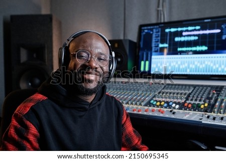 Portrait of cheerful African American music producer wearing headphones and eyeglasses sitting in recording studio smiling at camera