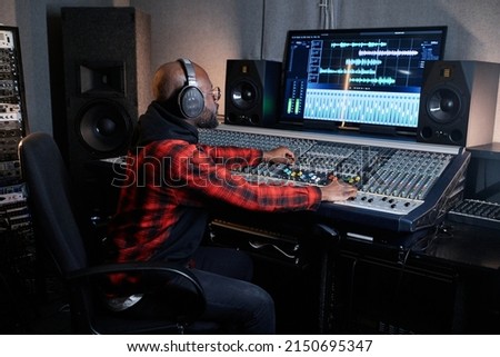 Professional African American recording producer wearing headphones sitting at mixing console working on music track