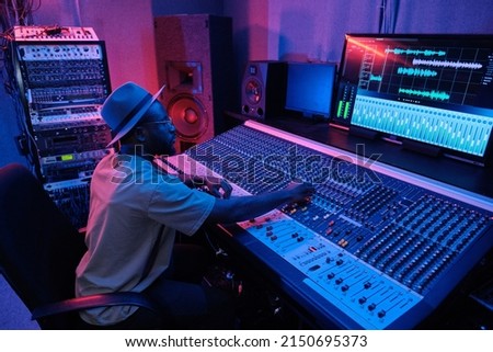 Stylish mature African American man wearing hat creating soundtrack using mixing console in recording studio in neon light Royalty-Free Stock Photo #2150695373
