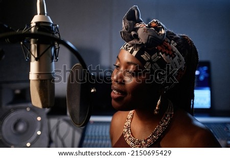 Portrait of stylish young African American female singer standing in front of microphone singing song in recording studio Royalty-Free Stock Photo #2150695429