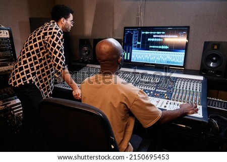 Mature African American producer and young singer creating music track using mixing console in recording studio