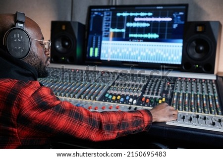 Modern African American sound engineer wearing headphones sitting at mixing console working on music track