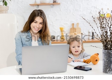 Young beautiful mother sitting at chair with little lovely daughter, having video call from family, smiling. Attractive woman with cute toddler using laptop, watching cartoons, concept of childhood.