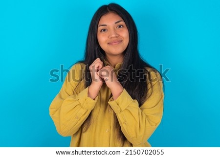 Charming serious young latin woman wearing yellow shirt over blue background  keeps hands near face smiles tenderly at camera