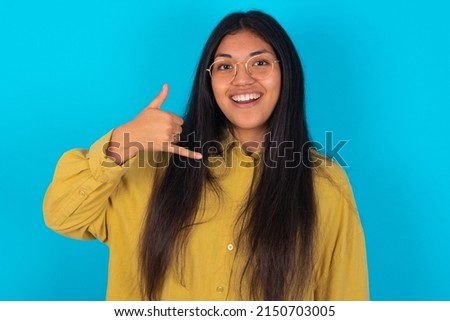 young latin woman wearing yellow shirt over blue background makes phone gesture, says call me back again, has glad expression.