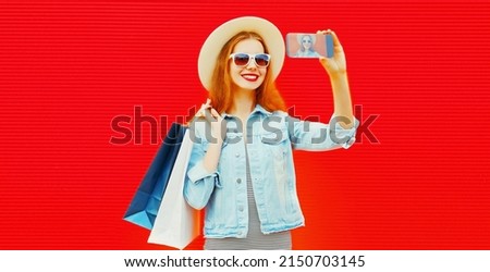 Portrait of happy smiling young woman with shopping bags taking selfie by smartphone wearing summer straw hat and denim jacket on red background