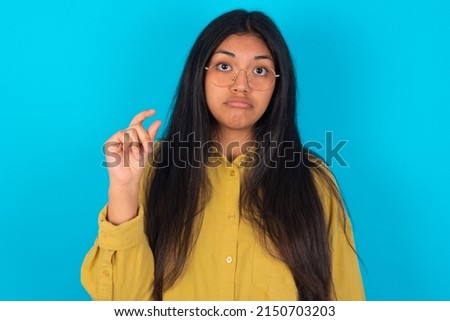 young latin woman wearing yellow shirt over blue background purses lip and gestures with hand, shows something very little.