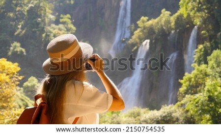 Millennial traveller or Inspiring travel blogger make photos for social media on beautiful tropical waterfall, amazing nature. Wanderlust traveling. Female traveler in trip in Southeast Asia Royalty-Free Stock Photo #2150754535