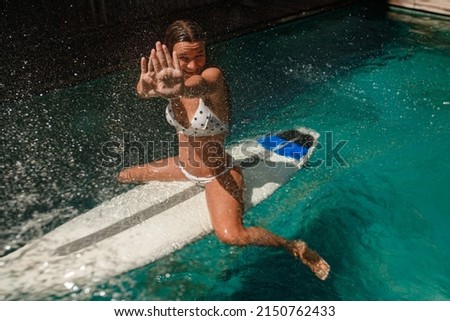 A tanned woman sits with a surfboard in the pool, covering her face from water drops, a rainbow is next to her, sunny weather