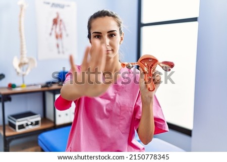 Young hispanic woman holding model of female genital organ at rehabilitation clinic with open hand doing stop sign with serious and confident expression, defense gesture 