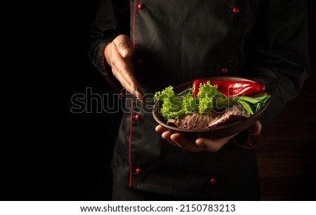 In the chef hand is a plate with sliced beef meat and vegetables. The concept of cooking on a dark background. European cuisine.