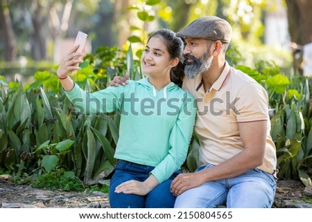Happy mature indian man and young teenager girl taking selfie with smart phone, smiling father and daughter using mobile phone outdoor in the park.