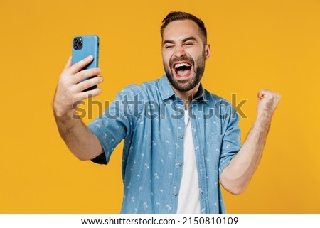 Young overjoyed cool happy man 20s in blue shirt white t-shirt doing selfie shot on mobile cell phone post photo on social network do winner gesture isolated on plain yellow background studio portrait