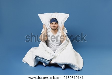 Full size body length happy young man in pajamas jam sleep mask rest relax at home cover ears with pillow sit wrap cover blanket duvet isolated on dark blue background Good mood night bedtime concept Royalty-Free Stock Photo #2150810161