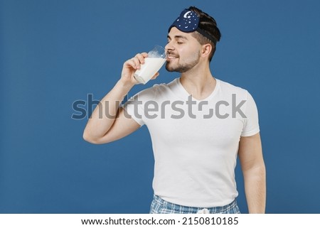 Young hungry fun calm happy caucasian man 20s wearing pajama jam sleep mask rest relax at home keep glass of milk drinking isolated on dark blue color background studio Good mood night bedtime concept