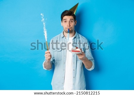 Funny guy celebrating birthday, holding b-day cake, firework and wearing party hat, standing over blue background