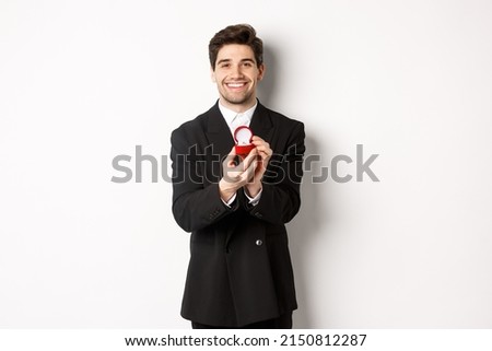Image of handsome boyfriend in black suit making a proposal, asking to marry him and showing wedding ring, standing over white background