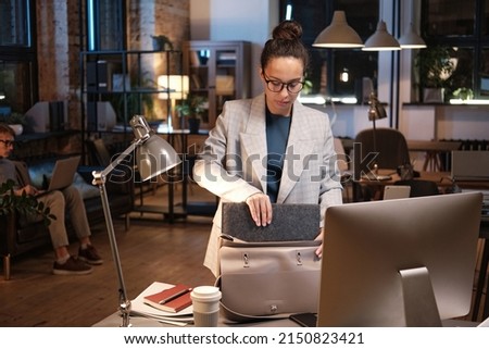 Serious young mixed race businesswoman in jacket putting folder in bag and taking work home while leaving office Royalty-Free Stock Photo #2150823421