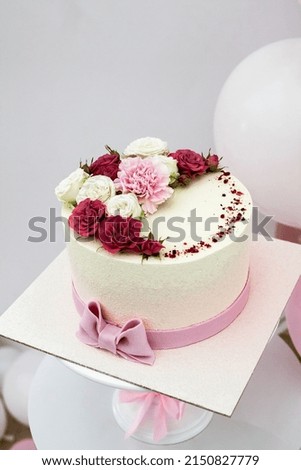 An appetizing festive yellow cake adorned with vibrant red, white and pink flowers sits on a white decorative stand in a room adorned with pink and white balloons. Royalty-Free Stock Photo #2150827779