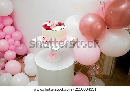 An appetizing festive yellow cake adorned with vibrant red, white and pink flowers sits on a white decorative stand in a room adorned with pink and white balloons. Royalty-Free Stock Photo #2150834331