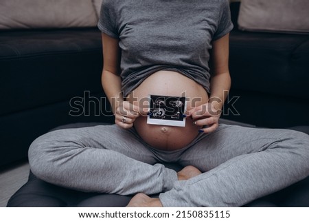 Close up cropped of pregnant woman in green dress showing ultrasound picture at camera, standing at home, young future mom demonstrating scan, waiting for first baby, pregnancy and motherhood concept