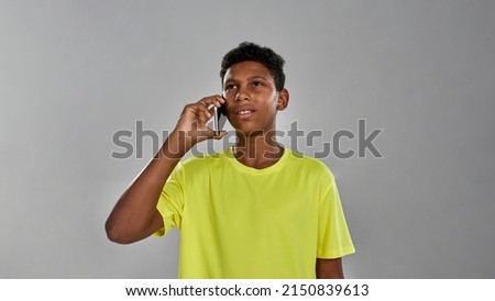 Thoughtful and focused sportive black boy talking on smartphone. Male youngster wearing yellow t-shirt. Concept of modern child lifestyle. Isolated on grey background. Studio shoot. Copy space