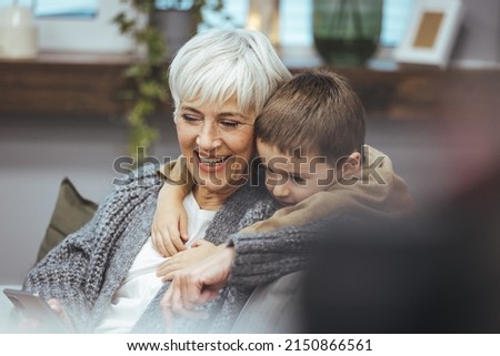 Grandma and grandson spend time together watching a funny video or cartoon on the phone, laughing while grandson hugs her. They enjoy their modern home.