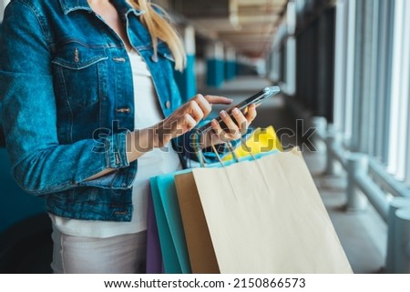 Cropped image of a young Caucasian woman writing a message or checking prices in stores on her mobile phone while standing with bags in the parking lot