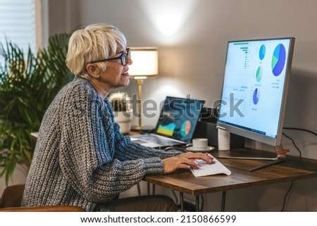  Senior stylish entrepreneur with notebook and pen wearing eyeglasses working on computer at home. Serious woman analyzing and managing domestic bills and home finance.