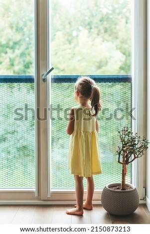Rear view of a little girl in yellow dress standing near the window and looking through it Royalty-Free Stock Photo #2150873213