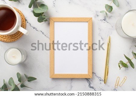 Top view photo of wooden photo frame candles cup of tea on rattan serving mat gold pen clips and eucalyptus on white marble background with empty space