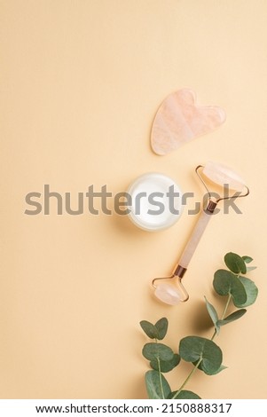 Beauty procedure concept. Top view vertical photo of rose quartz roller gua sha massager glass cream jar and eucalyptus branch on isolated pastel beige background