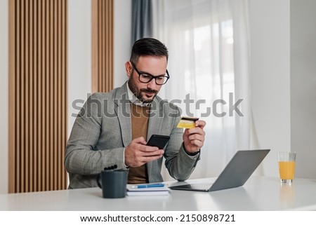 Businessman making card payment through smart phone. Serious Male professional using laptop at desk. He is wearing formals in office.